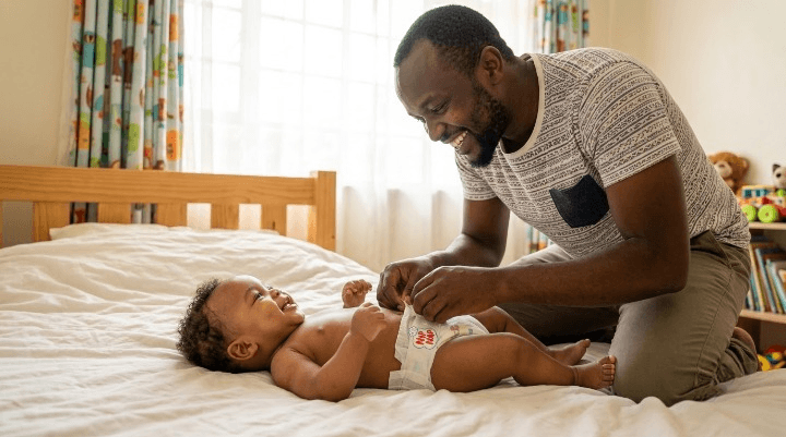 Kenyan father fastening a disposable nappy on a smiling baby lying on a bed
