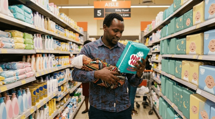 Parent holding a newborn and checking the label on a pack of baby nappies in a Kenyan supermarket aisle